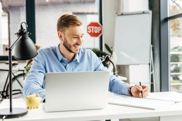 Giovane uomo d'affari sorridente usando il computer portatile e prendendo appunti sul posto di lavoro — Foto stock
