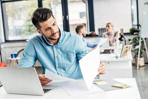 Konzentrierter Jungunternehmer arbeitet mit Papieren und Laptop im Büro — Stockfoto