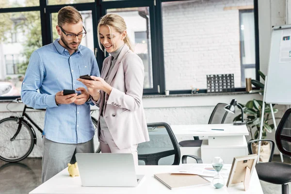 Lächelnde junge Geschäftskollegen, die Smartphones benutzen und im Büro reden — Stockfoto