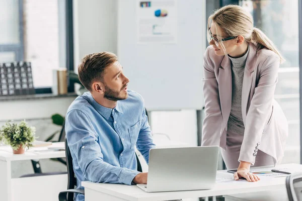 Jeune homme d'affaires et femme d'affaires parlant et se regardant tout en travaillant avec un ordinateur portable au bureau — Photo de stock