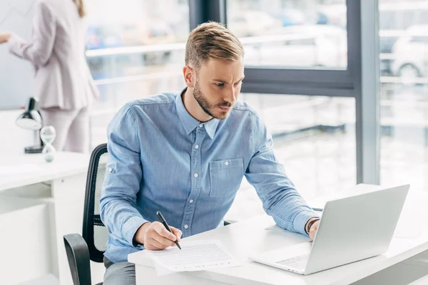 Joven empresario enfocado utilizando el ordenador portátil y tomando notas en el lugar de trabajo - foto de stock