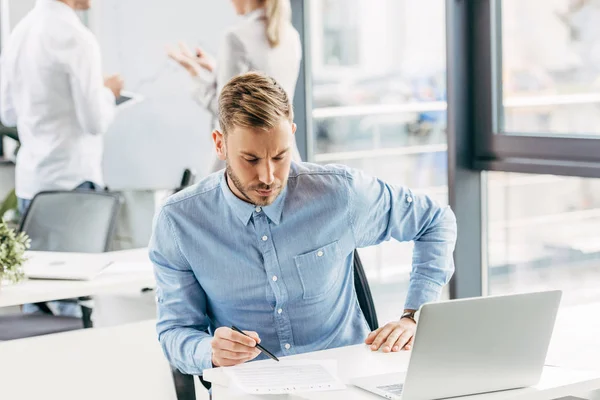 Joven hombre de negocios guapo que trabaja con el ordenador portátil y papeles en la oficina - foto de stock