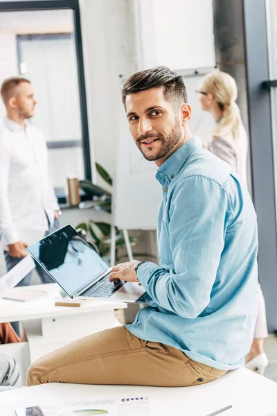 Jovem empresário bonito usando laptop e sorrindo para a câmera no escritório — Fotografia de Stock