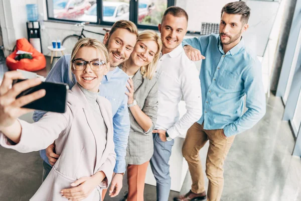 Vista de ángulo alto de equipo joven y feliz puesta en marcha tomando selfie con teléfono inteligente en la oficina - foto de stock