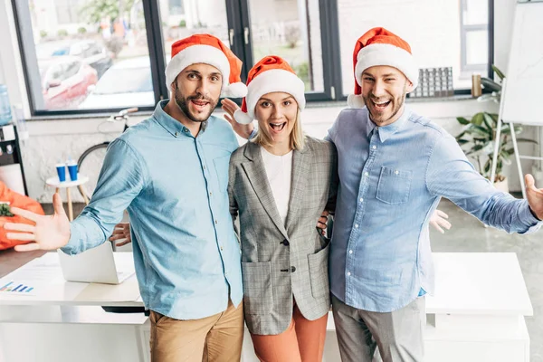 Fröhliches junges Unternehmerteam mit Weihnachtsmannhüten, das im Büro in die Kamera lächelt — Stockfoto