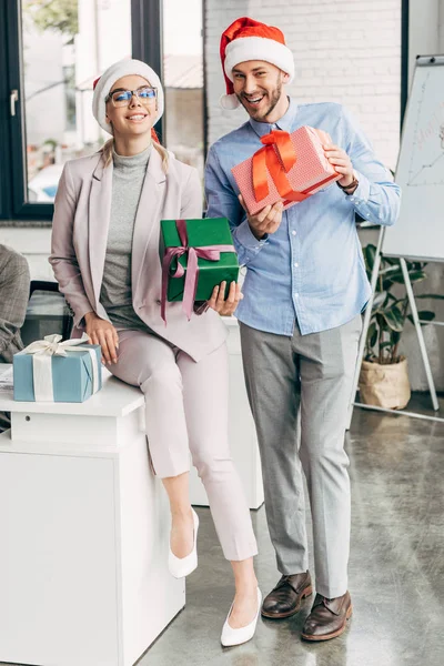 Feliz hombre de negocios y mujer de negocios en sombreros de santa celebración de cajas de regalo y sonriendo a la cámara en la oficina - foto de stock