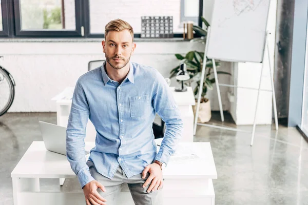 High angle view of handsome young businessman sitting on table and looking at camera in office — Stock Photo