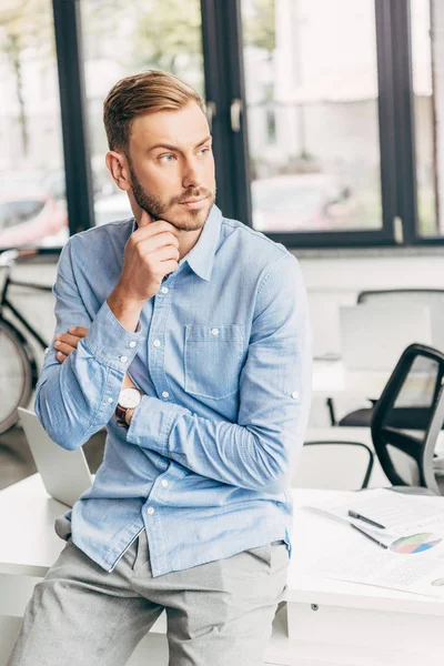 Pensativo joven hombre de negocios con la mano en la barbilla sentado en la mesa y mirando hacia otro lado en la oficina - foto de stock