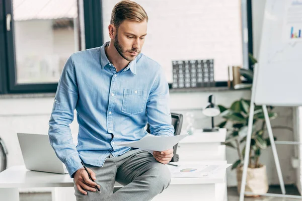 Serious young businessman holding papers and sitting on table in office — Stock Photo