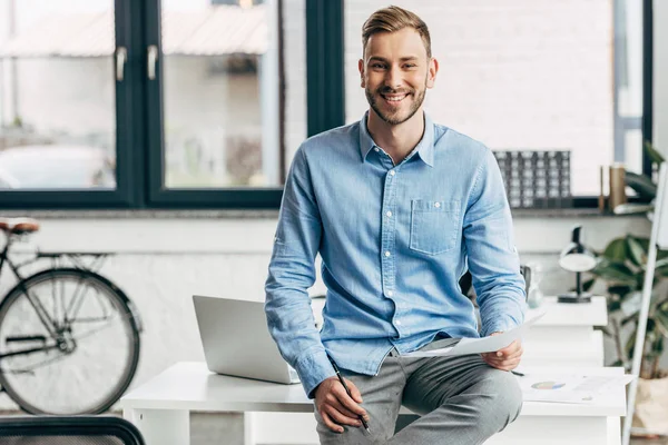 Handsome young businessman holding papers and smiling at camera in office — Stock Photo