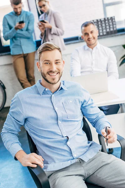 Schöner junger Geschäftsmann lächelt in die Kamera, während er mit Kollegen im Büro arbeitet — Stockfoto