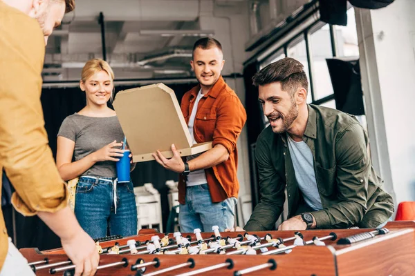 Gruppo di giovani uomini d'affari casuali che giocano a calcio balilla con la pizza in ufficio e si divertono insieme — Foto stock