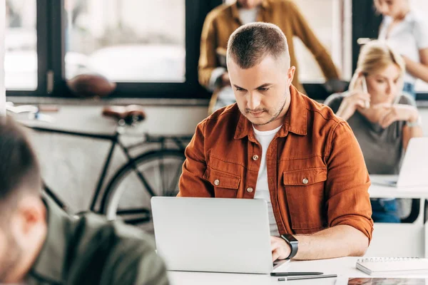 Attractive young man working with laptop at open space office with colleagues on background — Stock Photo