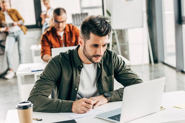 Nachdenklicher junger Mann arbeitet mit Laptop und Notizblock im Großraumbüro mit Kollegen im Hintergrund — Stockfoto