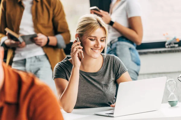 Sorrindo jovem que trabalha no escritório de espaço aberto com colegas em segundo plano e falando por telefone — Fotografia de Stock