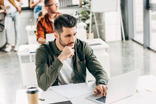 Joven hombre de negocios guapo que trabaja con el ordenador portátil con colegas borrosos en el fondo en la oficina - foto de stock