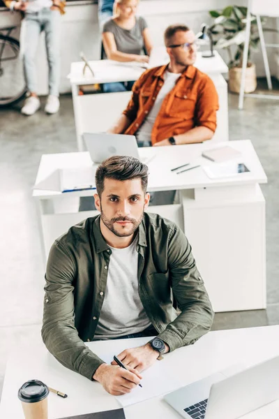 Hochwinkelaufnahme eines hübschen jungen Mannes, der im Großraumbüro arbeitet und mit Kollegen im Hintergrund in die Kamera schaut — Stockfoto