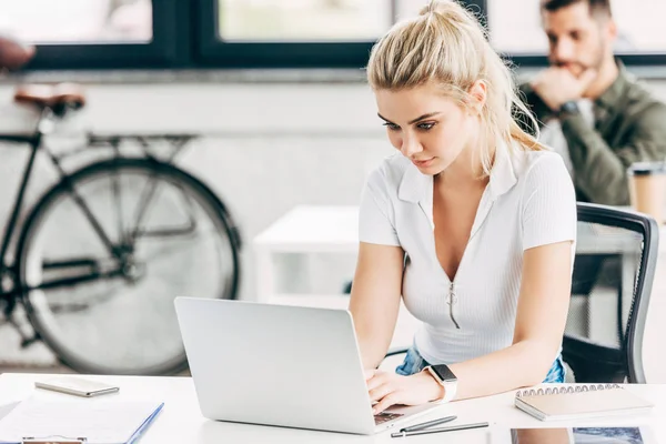 Attractive young woman working with laptop at office with colleague on background — Stock Photo