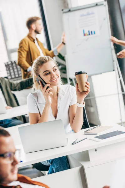 Glückliche junge Frau telefoniert und trinkt Kaffee im Büro mit Kollegen im Hintergrund — Stockfoto