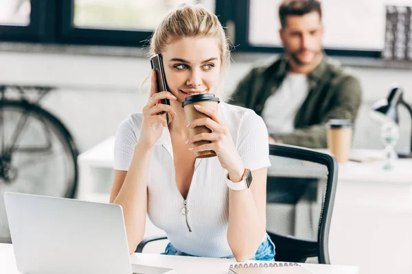 Красива молода жінка розмовляє по телефону і п'є каву в офісі з колегою на фоні — стокове фото