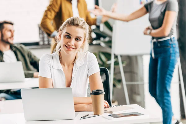 Belle jeune femme travaillant avec ordinateur portable et bloc-notes au bureau et regardant la caméra avec des collègues en arrière-plan — Photo de stock