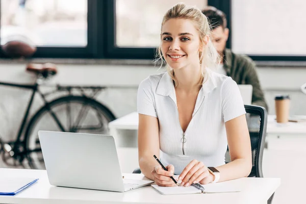 Hermosa mujer joven que trabaja con el ordenador portátil y bloc de notas en la oficina y mirando a la cámara - foto de stock