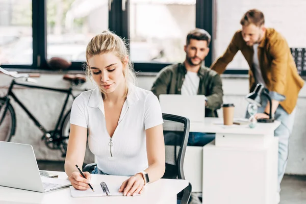 Beautiful young woman working with laptop and notepad at office with colleagues on background — Stock Photo