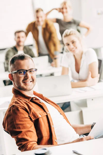Smiling young man working at open space office and looking at camera with colleagues on background — Stock Photo