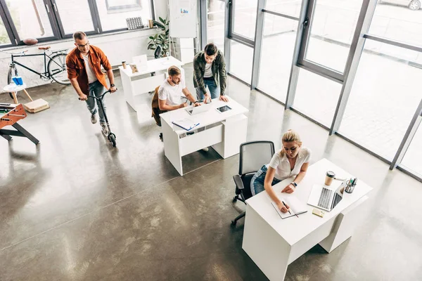 Blick aus der Vogelperspektive auf junge Unternehmer, die im modernen Großraumbüro gemeinsam an einem Start-up arbeiten, während der Mann Roller fährt — Stockfoto
