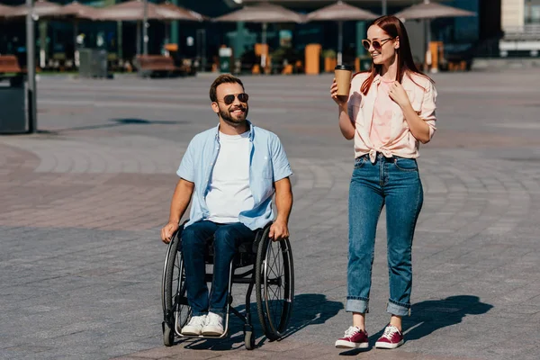 Guapo novio en silla de ruedas y novia con café en papel taza tener caminar en la calle - foto de stock