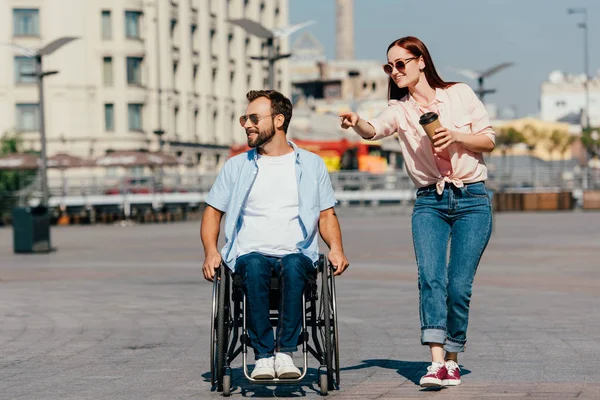 Atractiva novia apuntando en algo a guapo novio en silla de ruedas en la calle - foto de stock