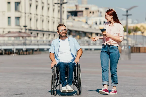 Sorrindo bonito namorado em cadeira de rodas e namorada com café para ir ter um passeio na rua — Fotografia de Stock