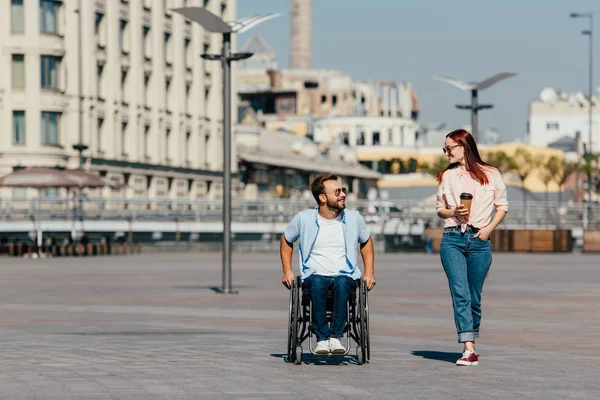 Guapo novio en silla de ruedas y novia con tener caminar en la ciudad y mirarse unos a otros - foto de stock