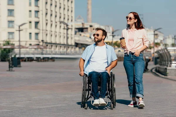 Bonito namorado em cadeira de rodas e namorada com café no copo de papel ter andar na rua e olhando para longe — Fotografia de Stock