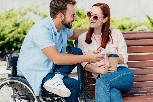 Novio guapo sonriente en silla de ruedas y novia sosteniendo el teléfono inteligente y mirándose en la calle - foto de stock
