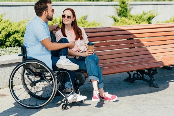 Novio guapo en silla de ruedas con teléfono inteligente y novia con café para ir a pasar tiempo en la calle - foto de stock