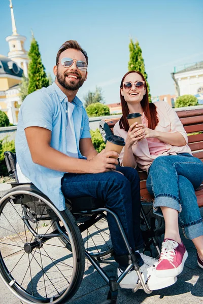 Смеющийся красивый парень в инвалидной коляске и девушка с кофе в бумажных стаканах на улице — стоковое фото