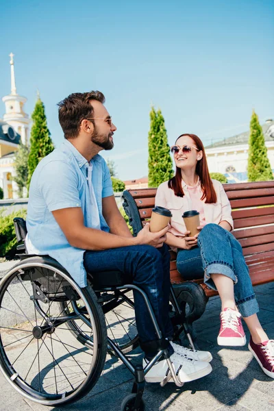 Novio alegre en silla de ruedas y novia con tazas de café desechables en la calle - foto de stock