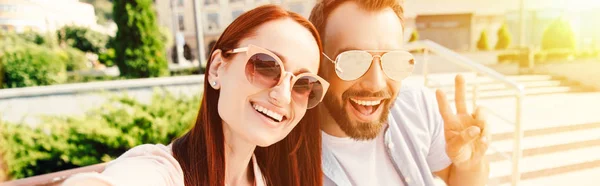 Vista panorámica de novio feliz y novia en gafas de sol mirando a la cámara en la ciudad, hombre mostrando señal de paz - foto de stock