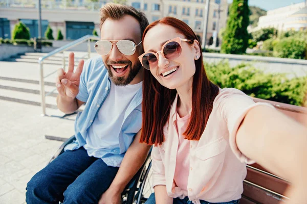 Punto de vista de la cámara de feliz novio en silla de ruedas y atractiva novia mirando a la cámara en la ciudad, hombre mostrando signo de paz - foto de stock