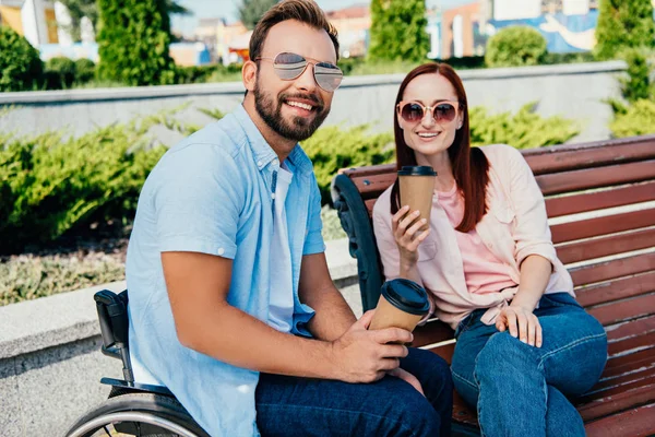 Sorrindo bonito namorado em cadeira de rodas e namorada olhando para a câmera e segurando copos de café descartáveis na rua — Fotografia de Stock