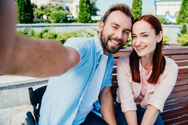 Camera point of view of smiling boyfriend in wheelchair and attractive girlfriend looking at camera in city — Stock Photo