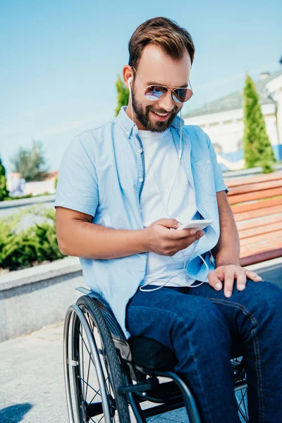 Smiling handsome man in wheelchair using smartphone and listening to music on street — Stock Photo