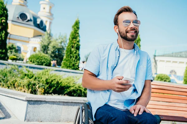 Hombre guapo sonriente en silla de ruedas sosteniendo el teléfono inteligente y escuchando música en la calle, mirando hacia otro lado - foto de stock