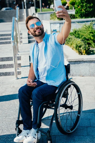 Bonito homem em cadeira de rodas tomando selfie com smartphone e ouvir música na rua — Fotografia de Stock