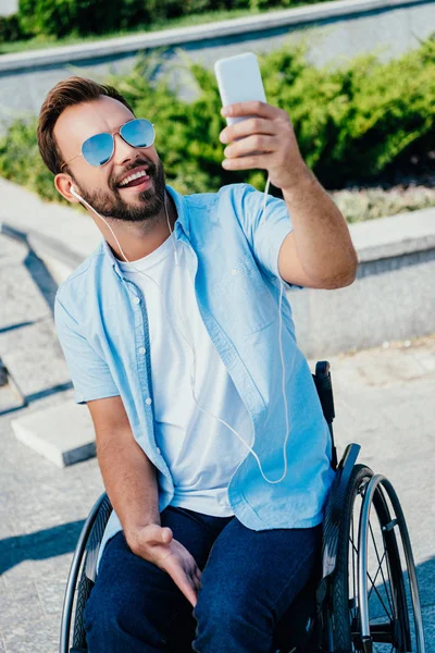 Homem bonito em cadeira de rodas colando a língua para fora e tomando selfie com smartphone na rua — Fotografia de Stock