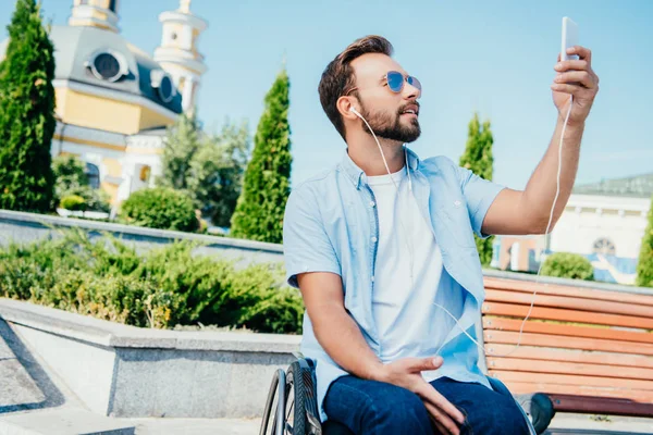 Handsome man in wheelchair taking selfie with smartphone and listening to music on street — Stock Photo