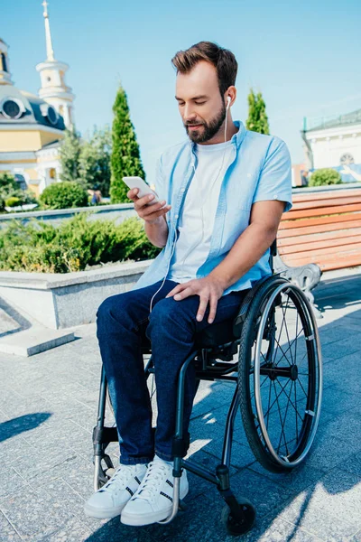 Handsome man in wheelchair using smartphone and listening to music on street — Stock Photo