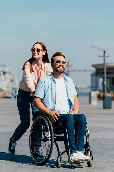 Attractive girlfriend pushing handsome disabled boyfriend in wheelchair on street — Stock Photo