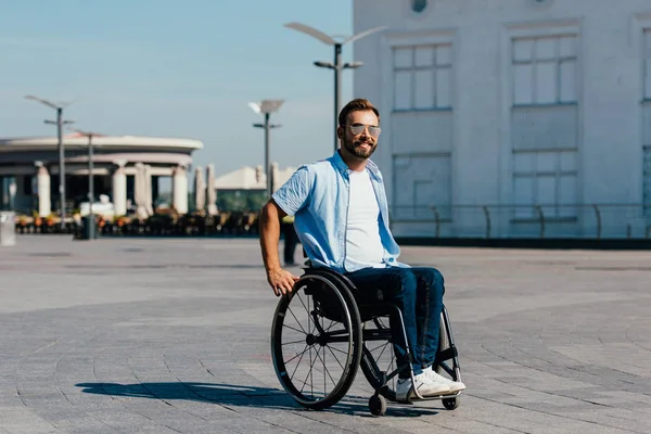 Sonriente discapacitado guapo hombre en gafas de sol usando silla de ruedas en la calle - foto de stock